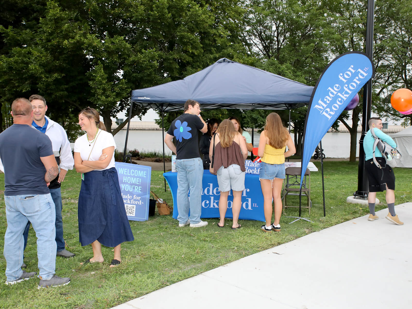 People stand and talk near a “Made for Rockford” booth with a blue tent at an outdoor event on grass, with trees and a sidewalk in the background.