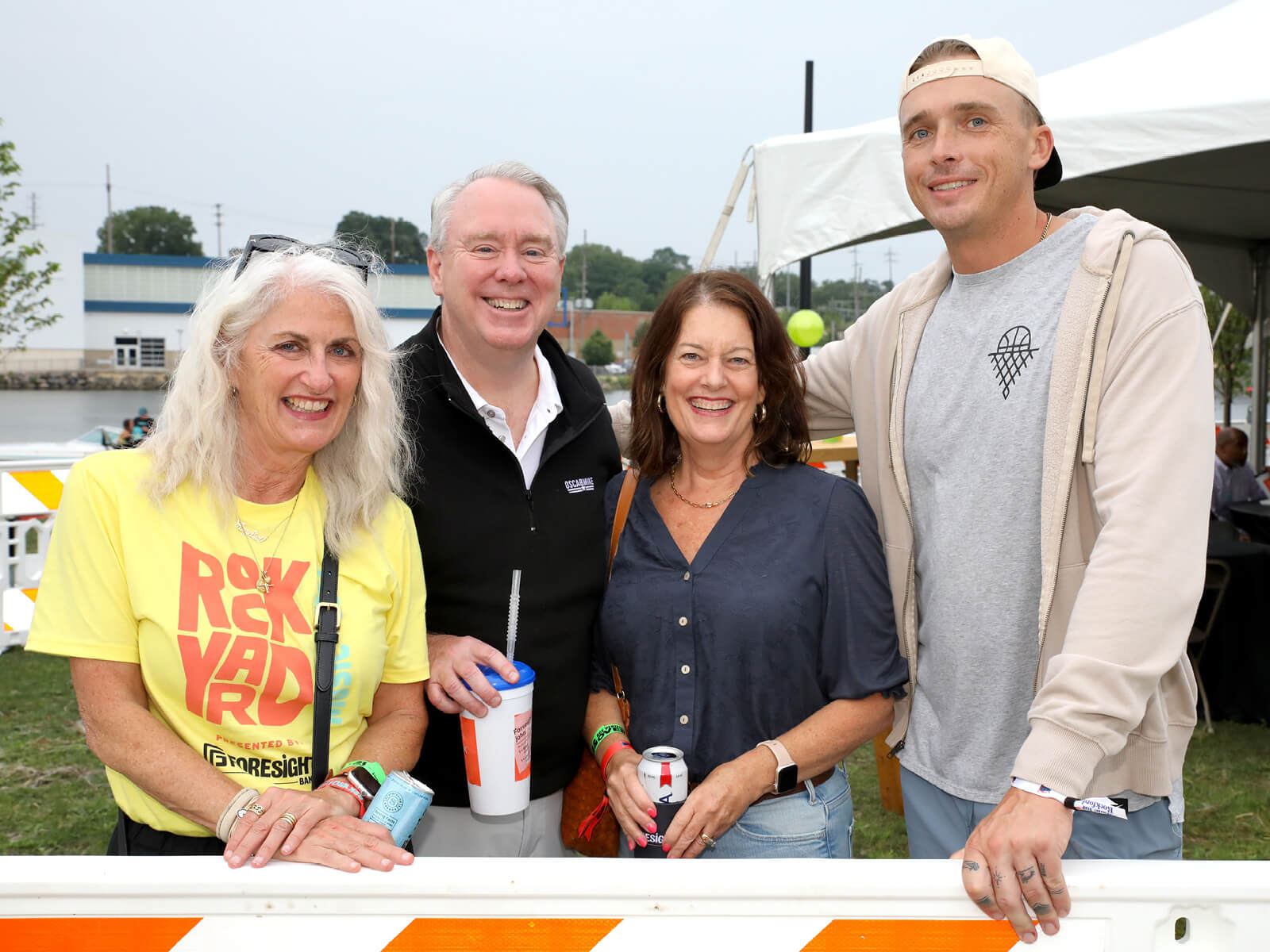 Four smiling adults pose at an outdoor event: a woman in a yellow RockYard shirt, two guests with drinks, and a man in a cap.