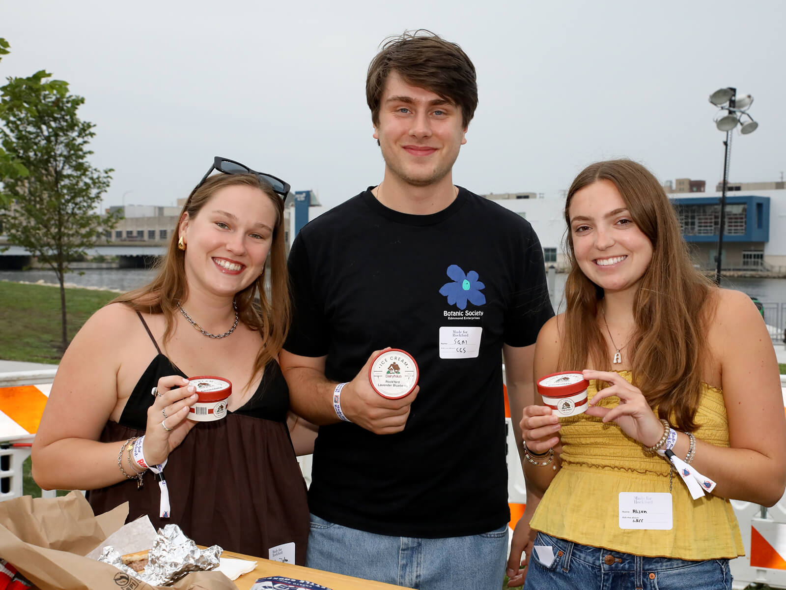 Three smiling young adults hold small dessert cups at an outdoor event, wearing name badges; a cheerful, friendly festival atmosphere.