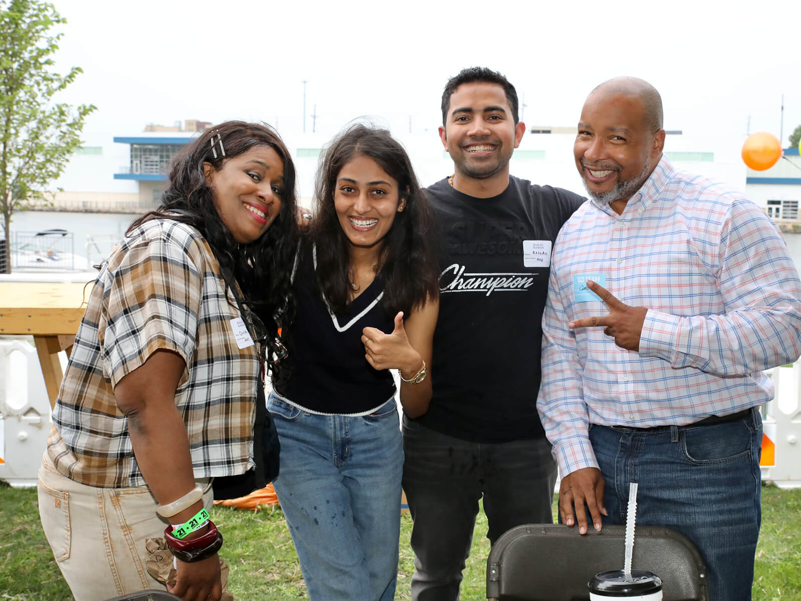 Four diverse friends smile and pose outdoors at a sunny community event; two give thumbs-up and peace signs, conveying joy and camaraderie.