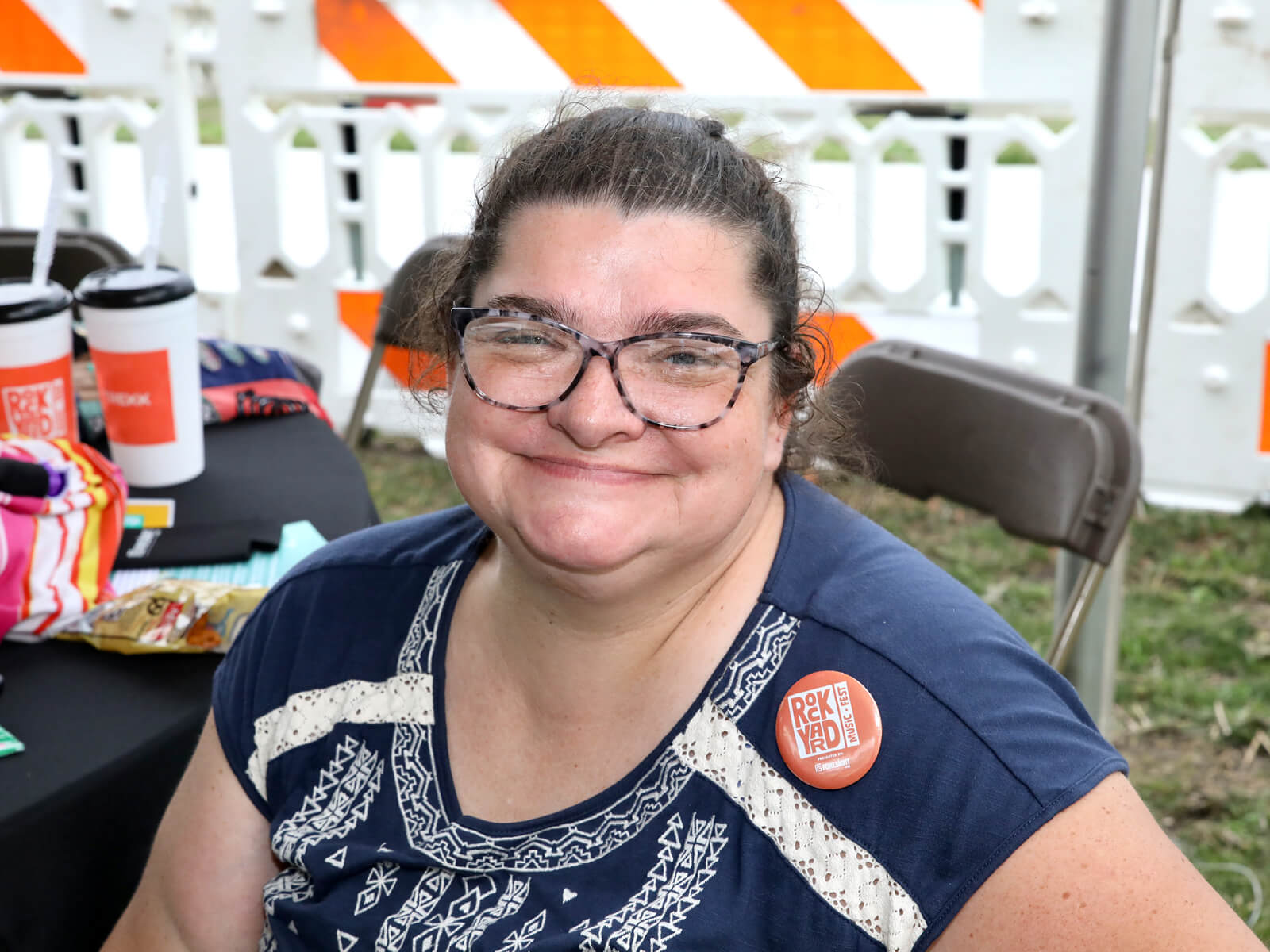 Smiling woman wearing glasses and a patterned navy shirt with a “RockYard” button, seated at an outdoor event table.