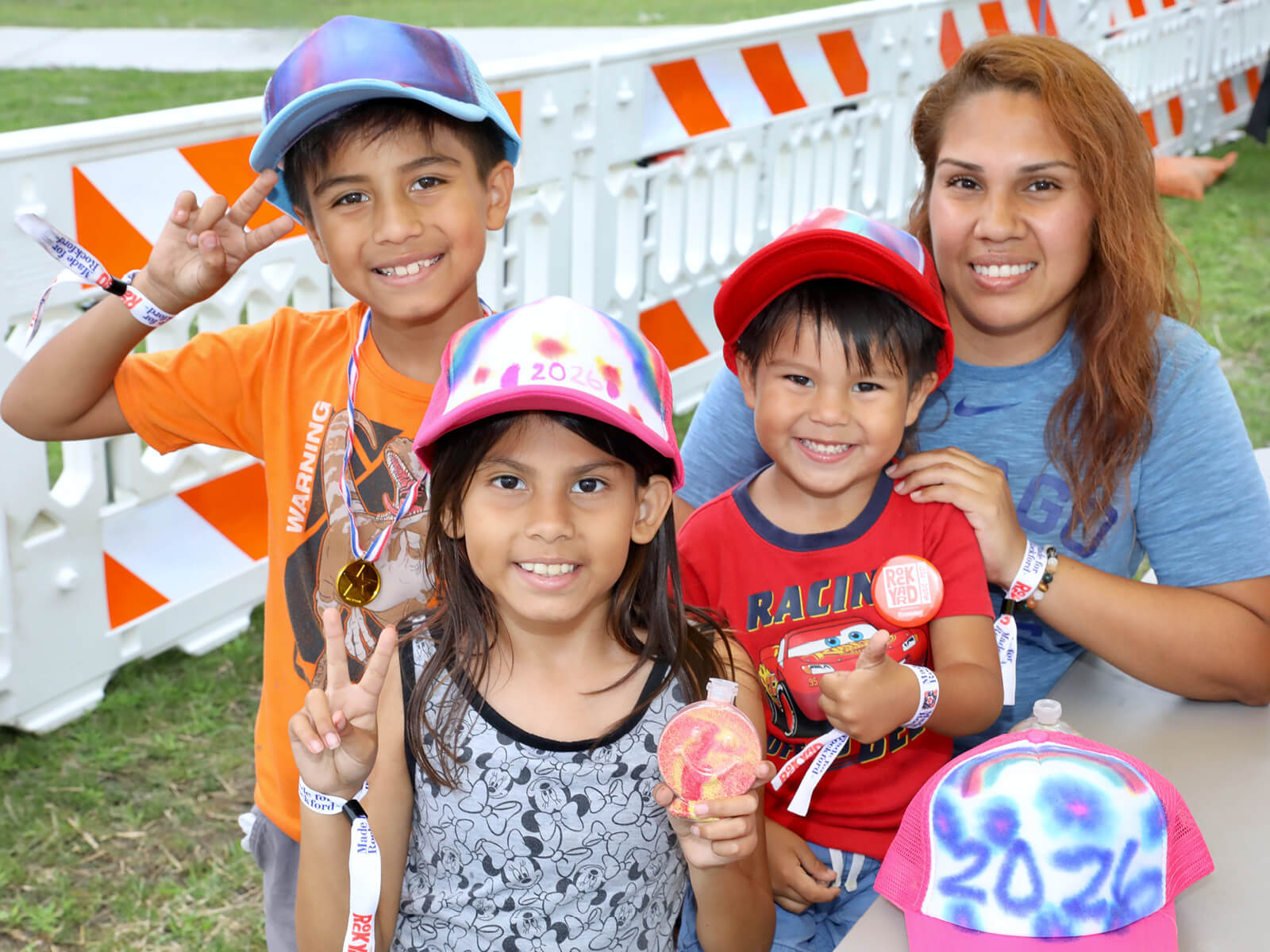 Smiling family at RockYard Music Fest: children pose with medals and colorful caps as their mother stands behind them near safety barriers.