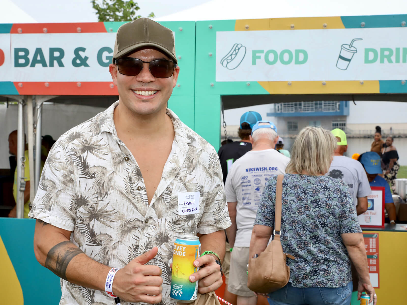 Smiling man in sunglasses and floral shirt holds a drink at a colorful outdoor RockYard Music Fest, with crowd behind him.