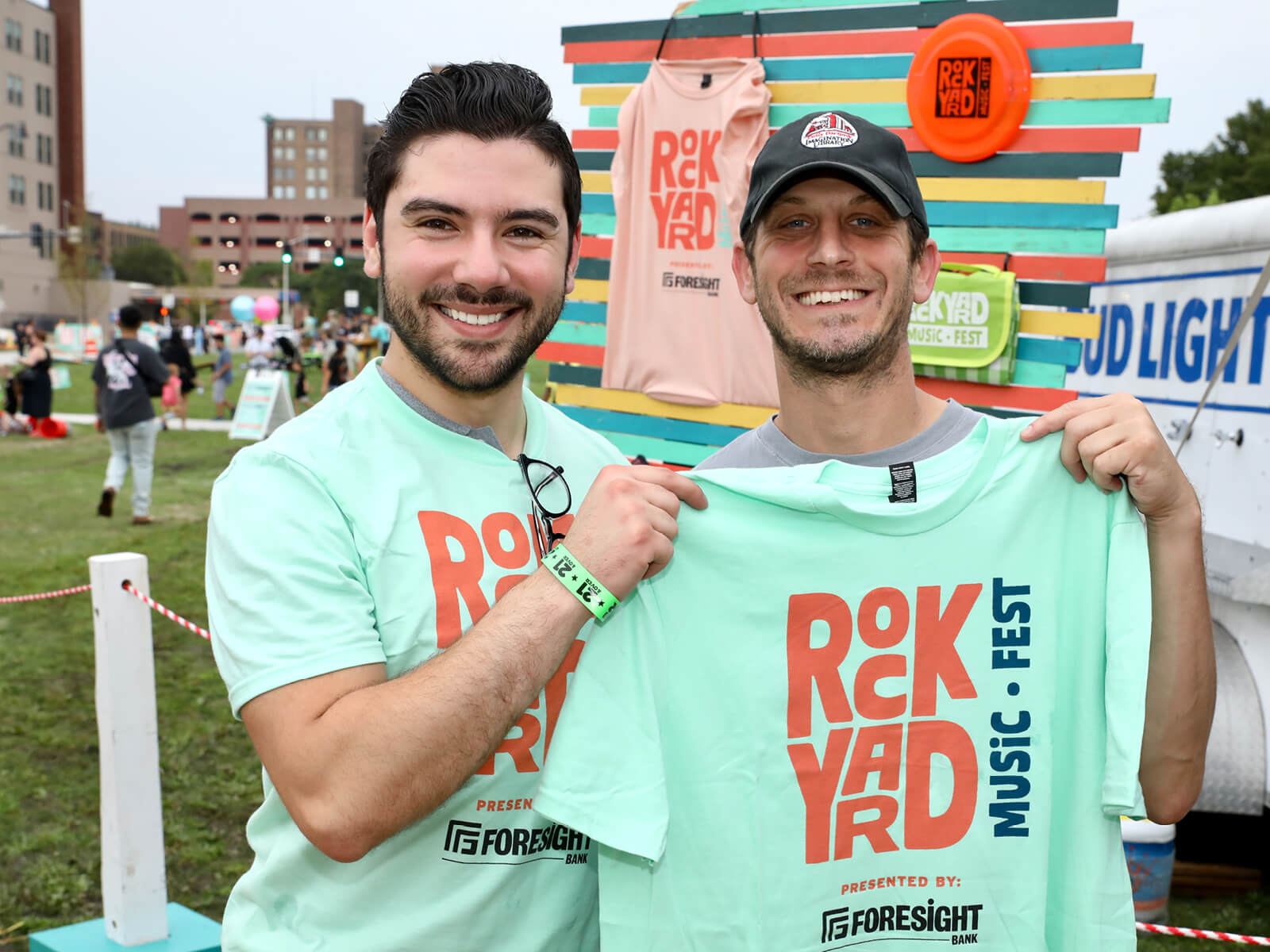 Two smiling men hold up matching “RockYard Music Fest” T-shirts at an outdoor festival, with colorful decor and a lively crowd behind them.