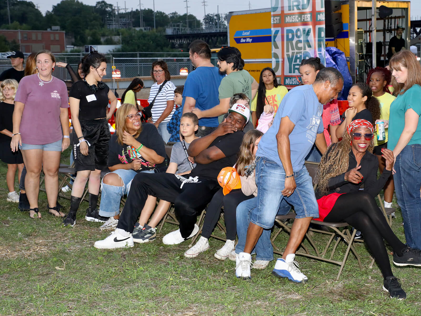 Diverse crowd gathers outdoors at a lively community event, with people seated and standing near a colorful music sign, smiling and playing a game of musical chairs.