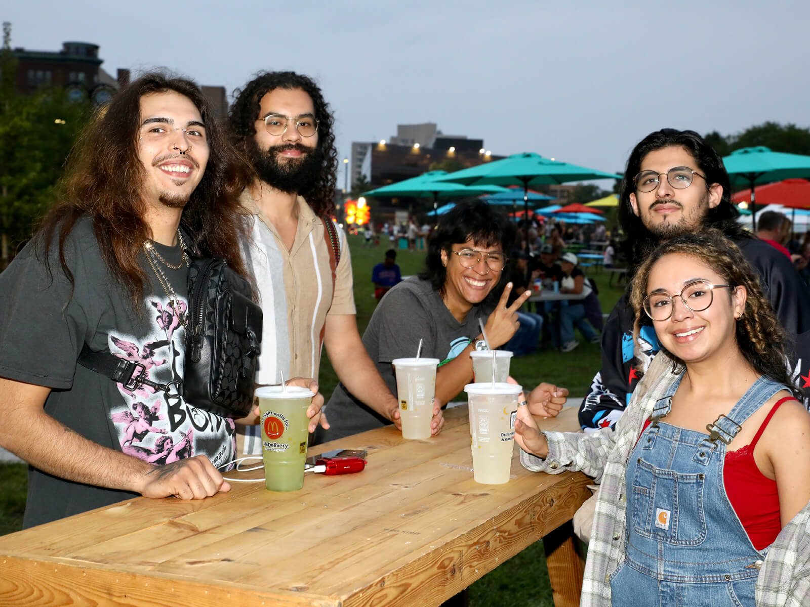 Five friends smile around a wooden table at RockYard Music Fest, holding drinks; one flashes a peace sign beneath colorful umbrellas.