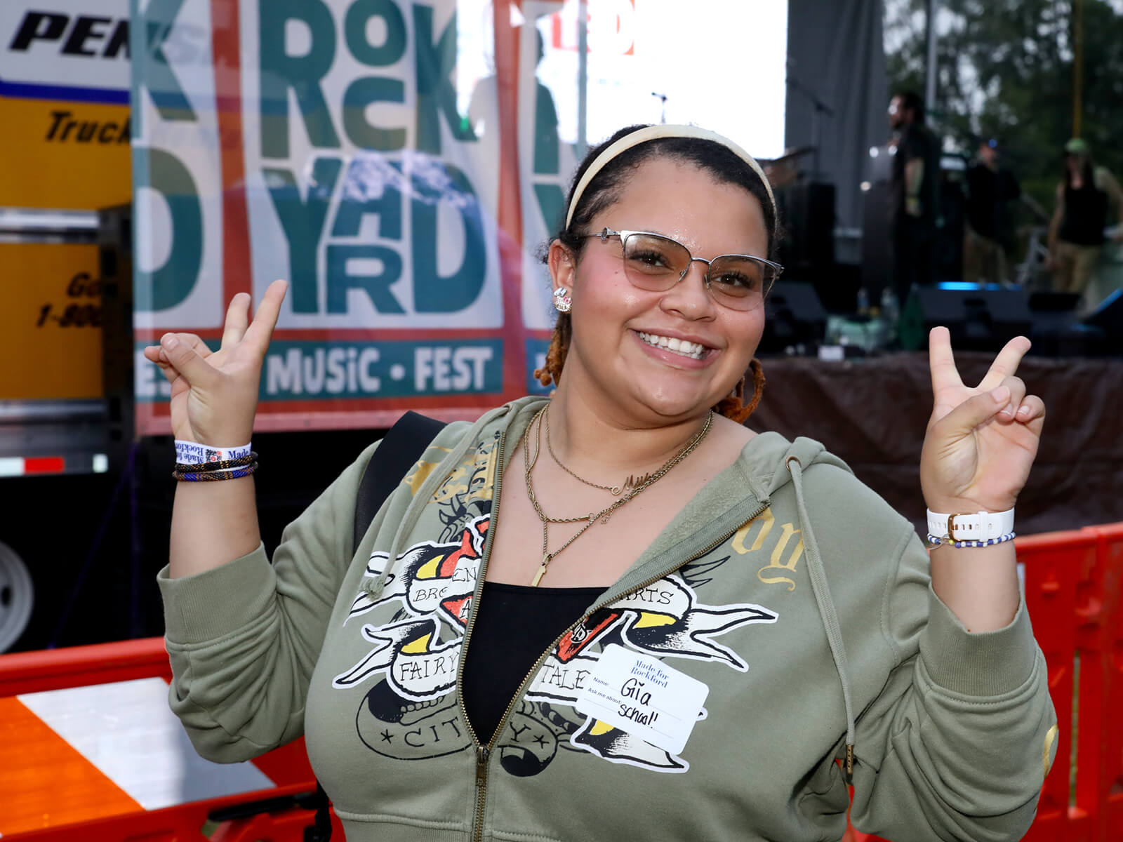 Smiling woman in glasses poses with both hands raised in peace signs at RockYard Music Fest, with a colorful event banner behind her.