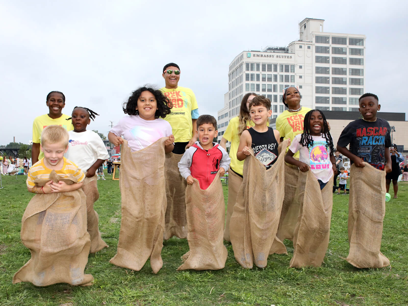 A group of children participate in a sack race on a grassy field with a tall building in the background.