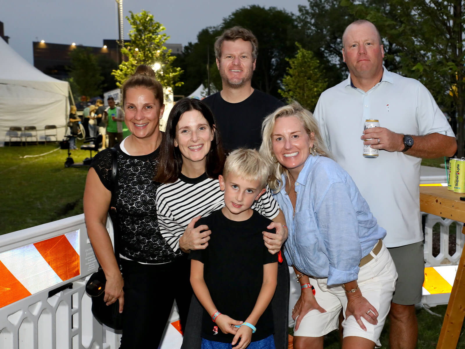 Six adults and a boy smile for a group photo at RockYard Music Fest, with tents, trees, string lights, and barricades behind them.