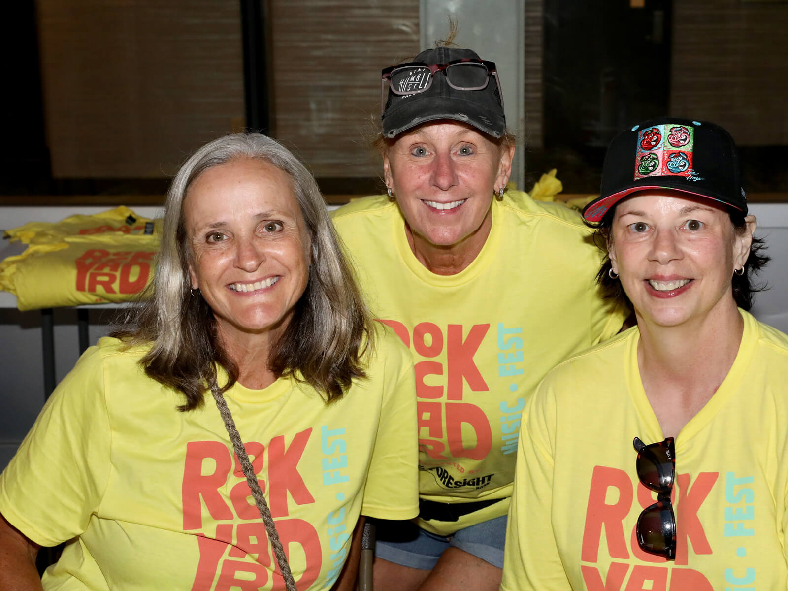 Three smiling women in matching yellow graphic shirts pose indoors; two wear hats and one has sunglasses, creating a cheerful event mood.