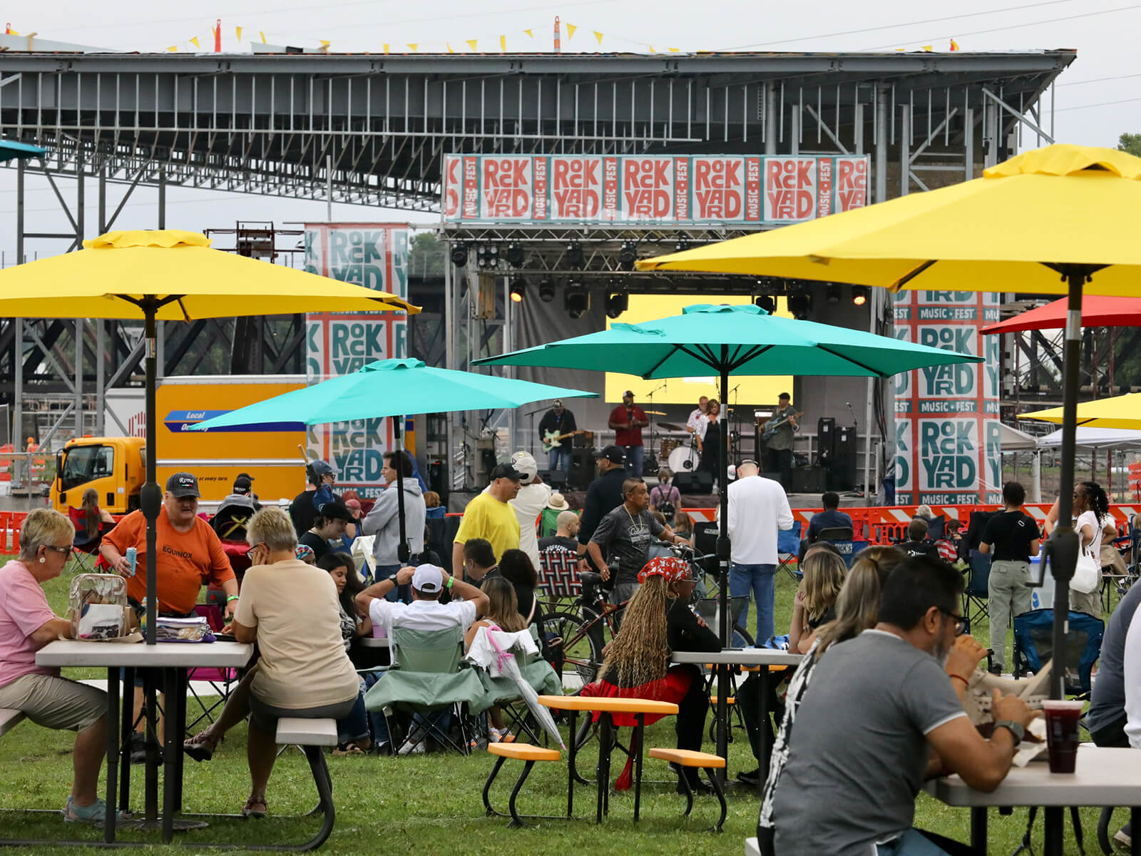 Crowded outdoor RockYard Music Fest with colorful umbrellas, picnic tables, food trucks, and a stage decorated with bold graphic panels; lively mood.