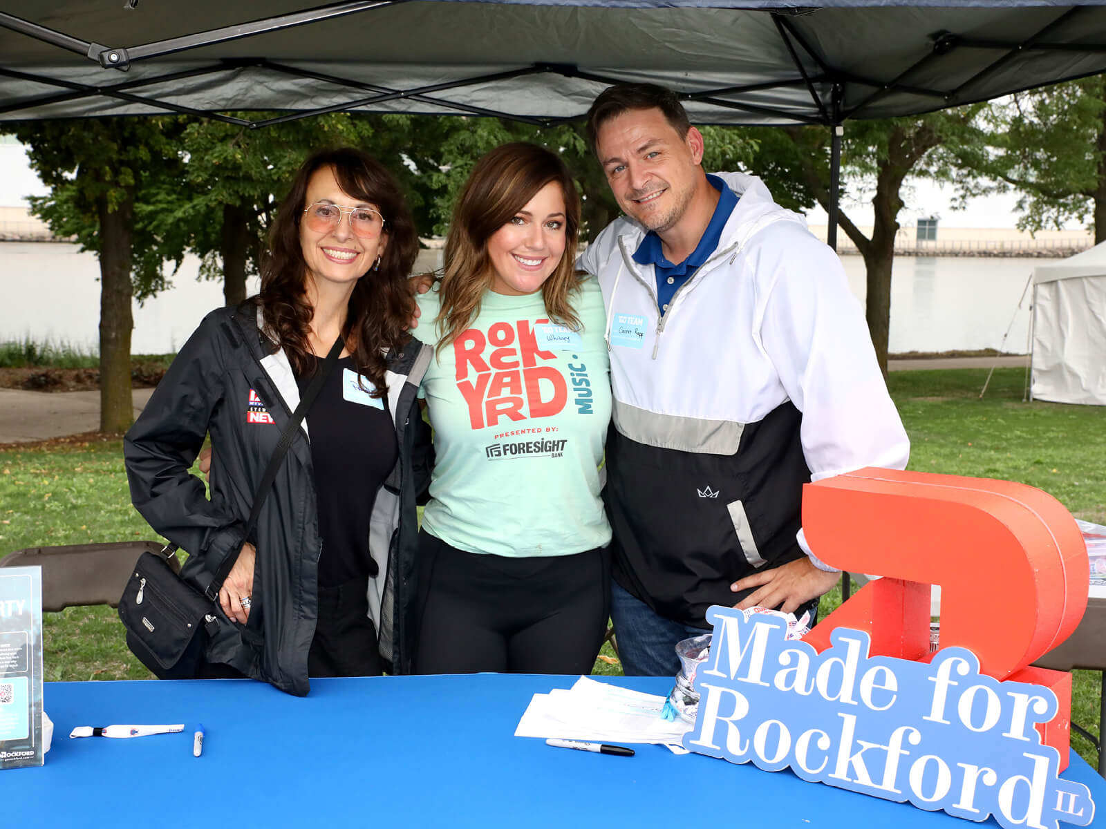 Three smiling adults pose at a “Made for Rockford” community event booth beneath a canopy, with brochures and a blue tablecloth.