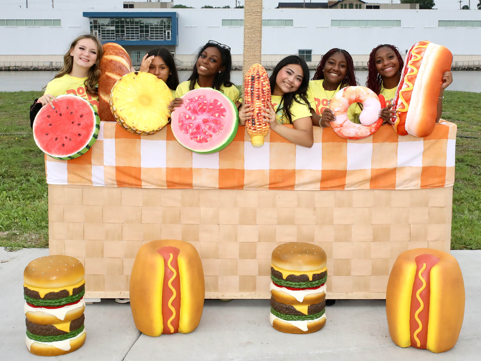 Smiling women pose at a playful food booth with giant fruit slices, burgers, and hot dogs, creating a bright, festive outdoor scene.