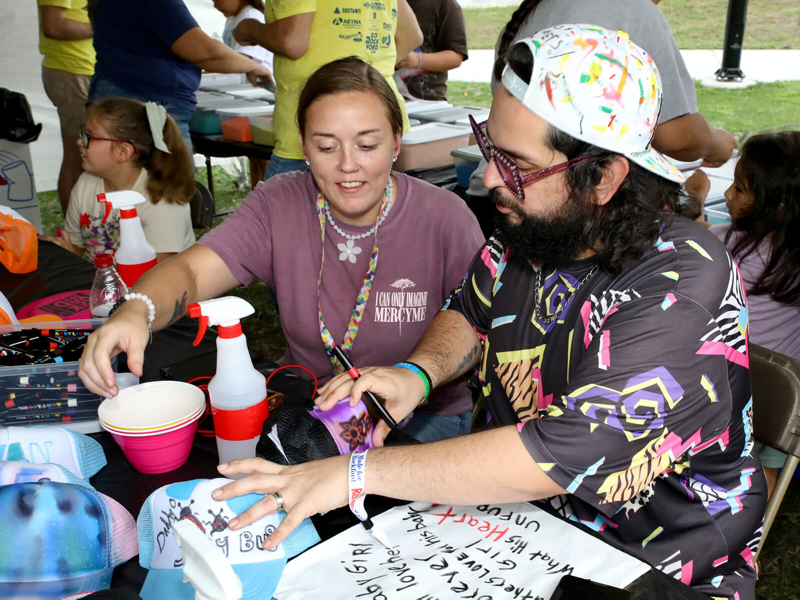 At an outdoor community event, a woman and bearded man in colorful attire collaborate on a painted craft at a busy table.