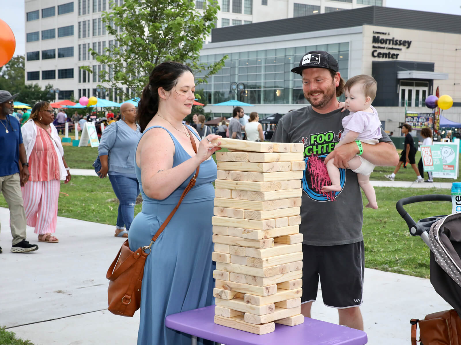 At a lively outdoor festival, a woman and smiling father holding a baby play giant Jenga, with the Morrison Center and crowd behind them.