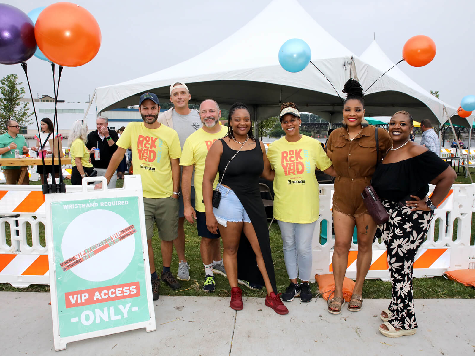 Diverse group smiles at a community event beneath a white tent, surrounded by colorful balloons and a “VIP Access Only” sign.