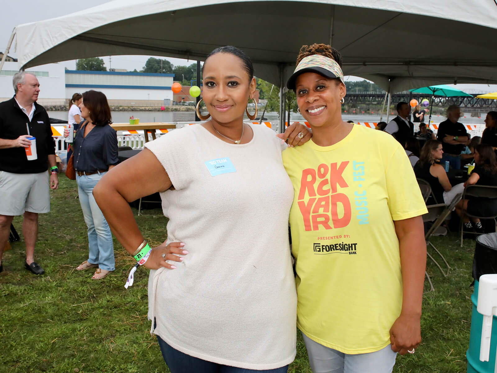 Two smiling women pose together at an outdoor community event under a tent, wearing wristbands and casual shirts, conveying warmth and friendship.