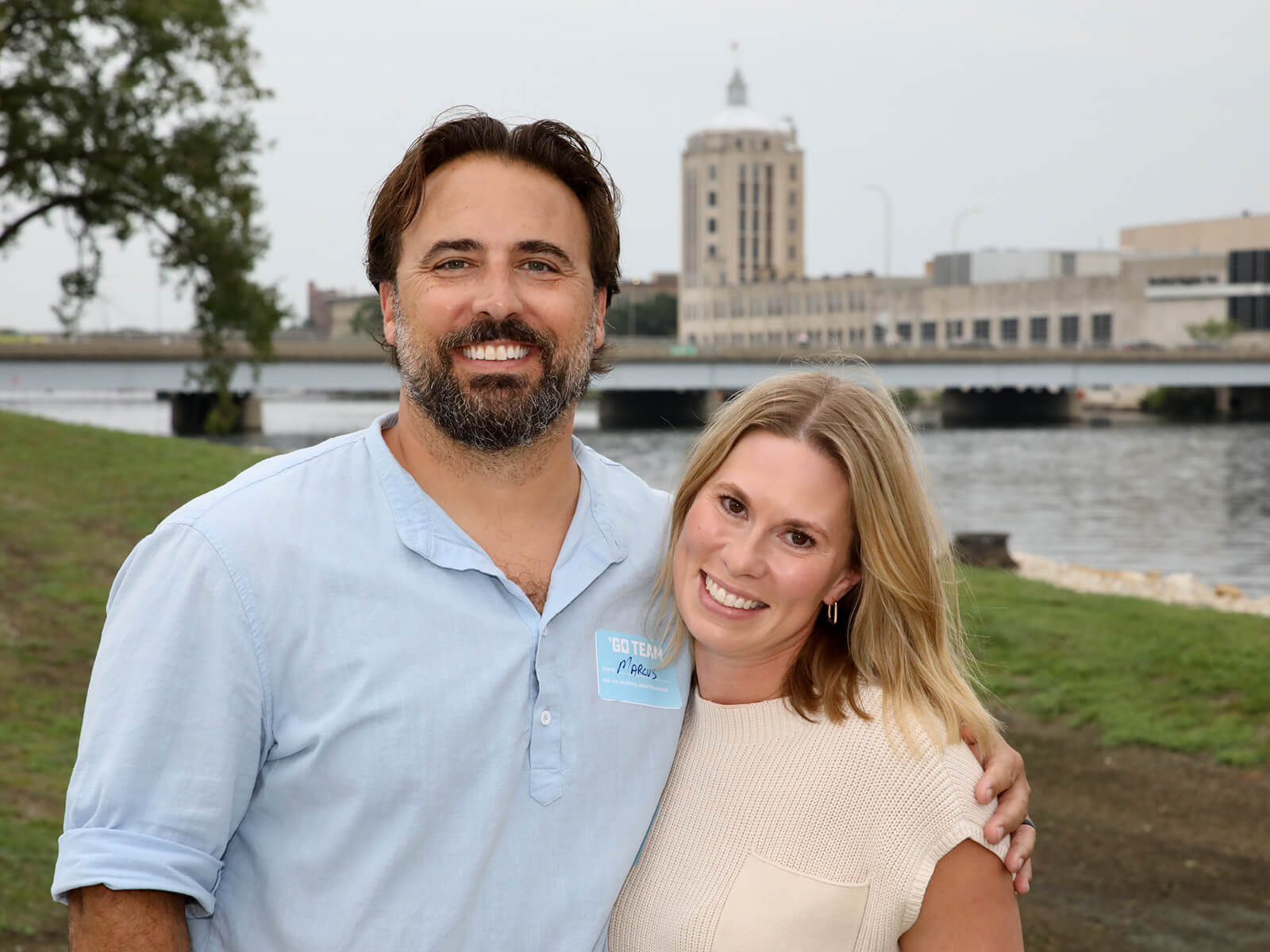 Smiling couple poses by a riverside, with a bridge and city tower behind them; their relaxed expressions convey warmth and happiness.